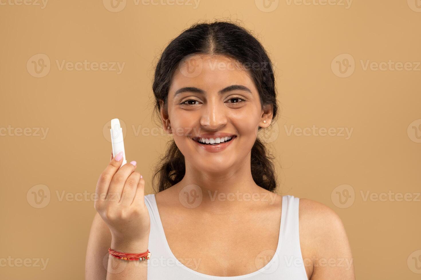 A young Indian woman is happily showcasing a skincare product while pampering herself. She exudes confidence and beauty against a warm, neutral backdrop that highlights her glowing skin. photo