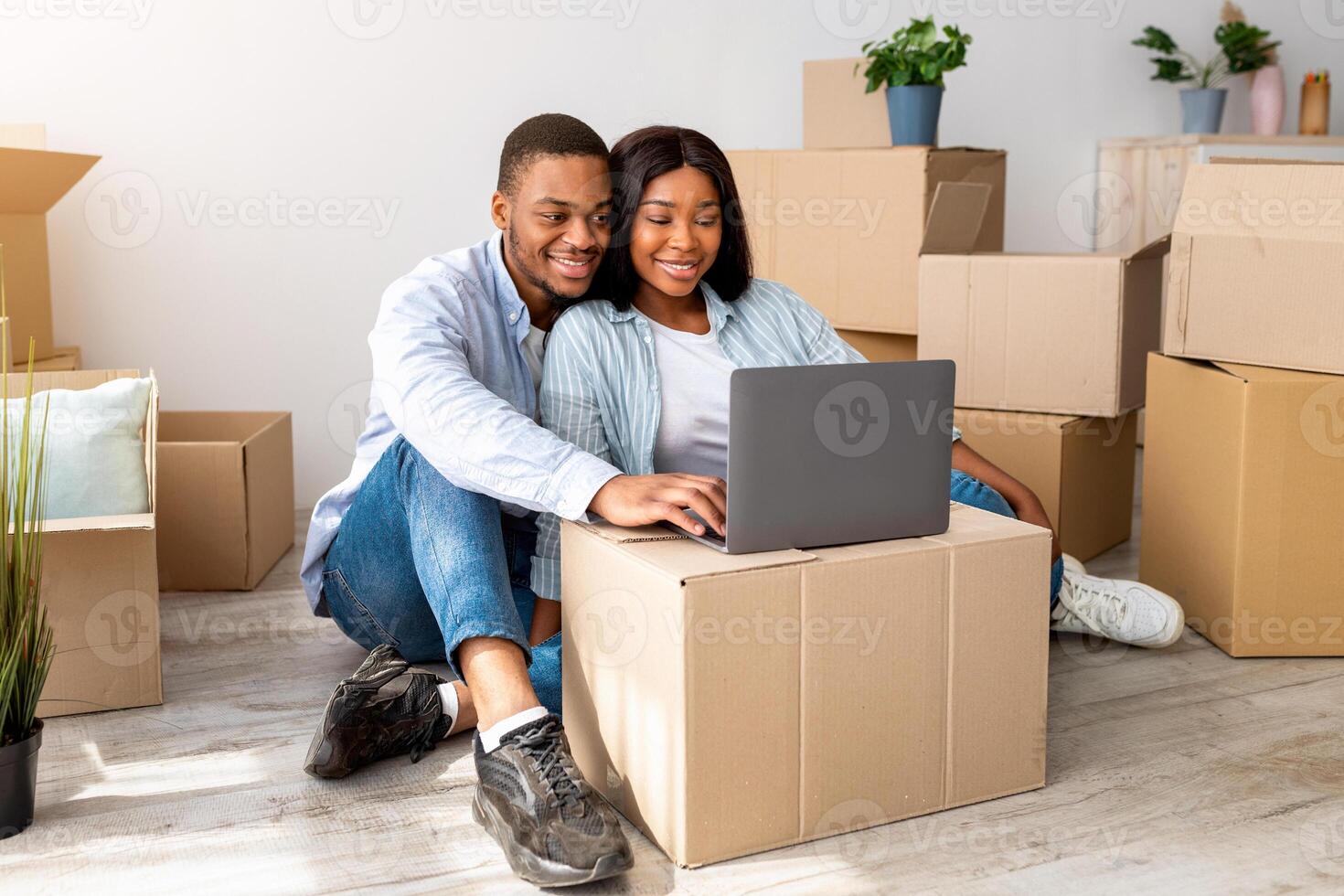 Happy african american spouses having call via laptop, sitting among carton boxes in new apartment and smiling to computer webcamera. Black couple telling news about relocation to family photo