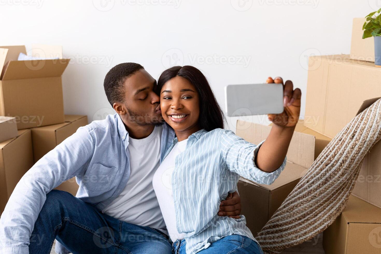 Happy black spouses taking selfie in their new apartment, celebrating moving day, sitting on the floor near carboard boxes. Loving husband kissing wife in cheek photo