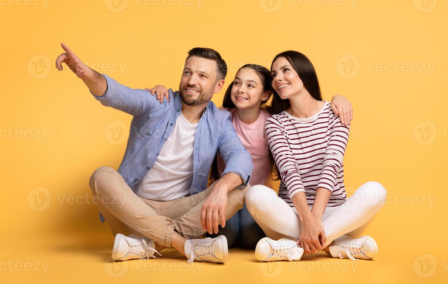 A father, mother, and their daughter sit together on the floor, showing affection. The father points excitedly, while the mother and daughter share smiles. photo