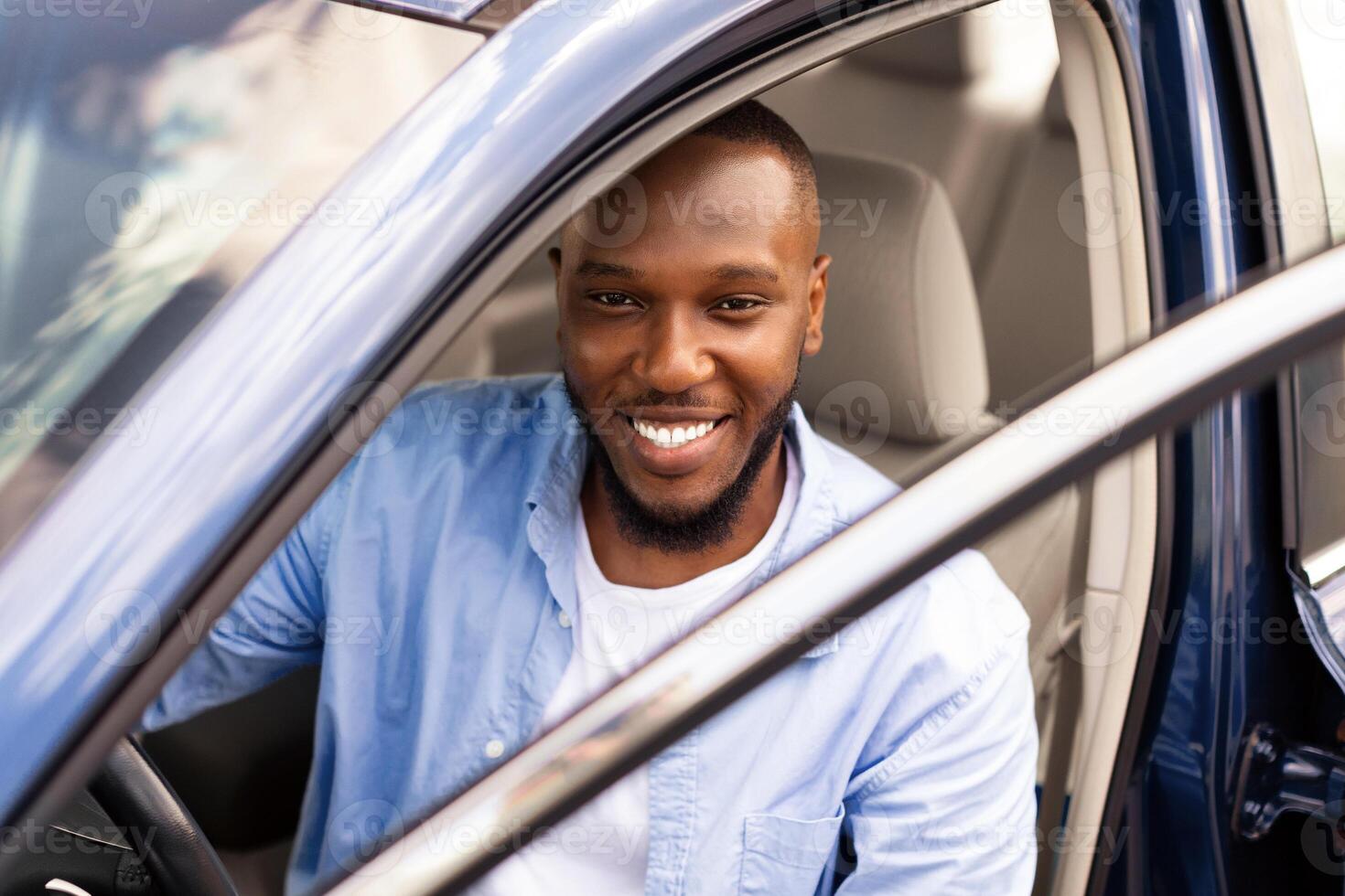 Positive young African American guy sitting on driver's seat. Happy black male posing looking at camera through open automobile door and smiling. Auto test-drive, leasing, buying, rental concept photo