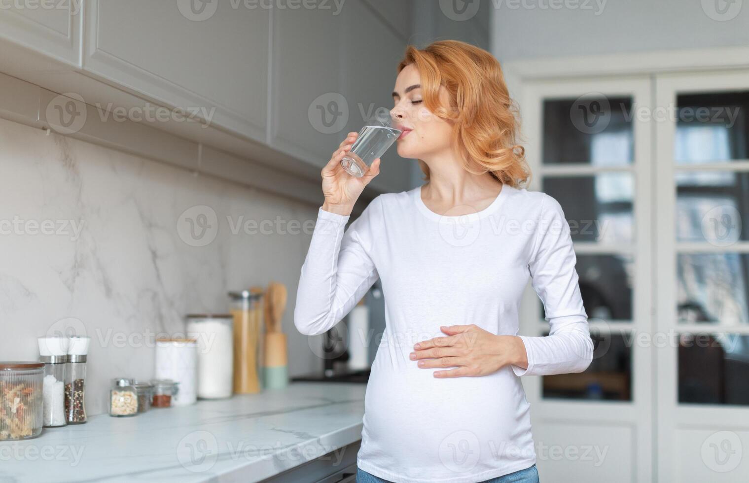 A young pregnant woman stands in a bright kitchen, sipping water from a glass. She gently rests her hand on her baby bump, enjoying her morning routine in a calming environment. photo
