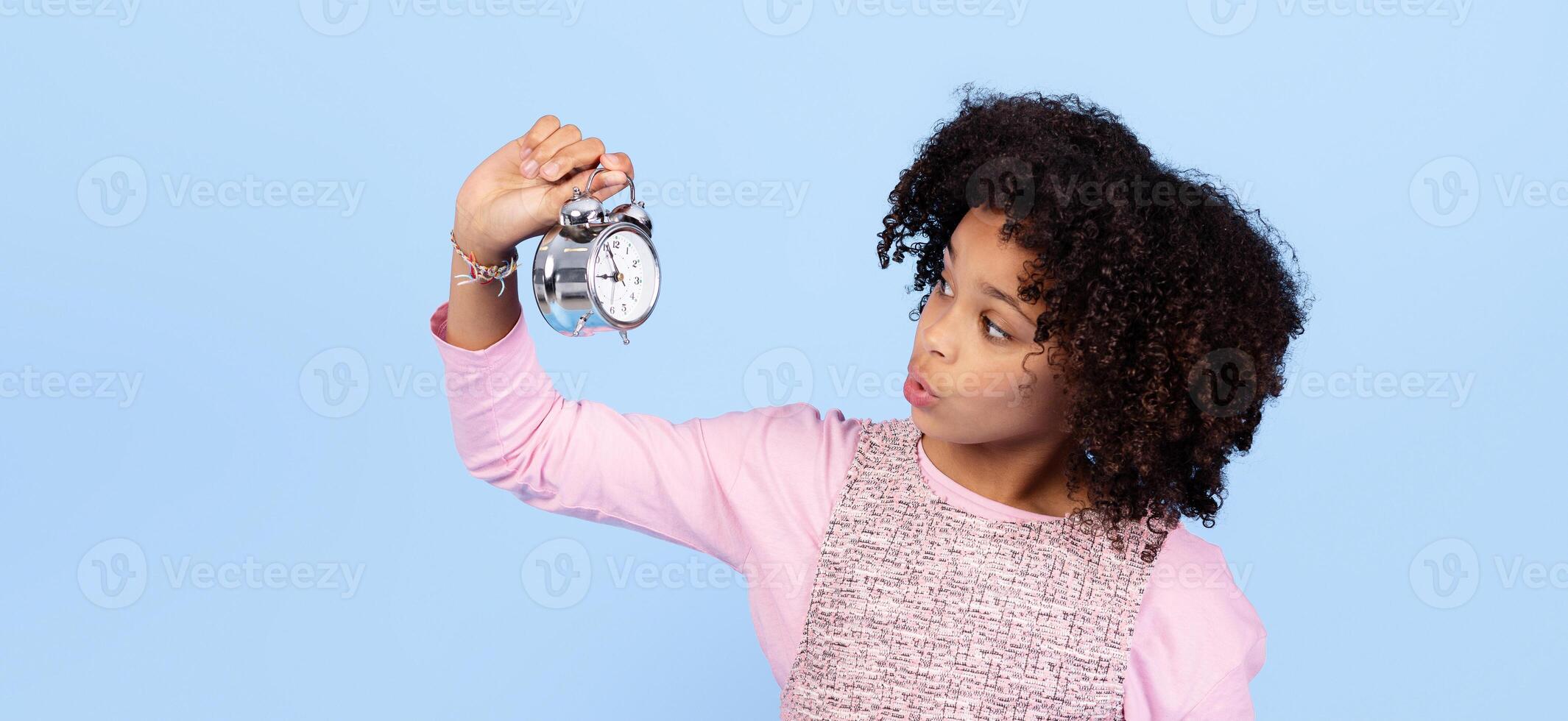 Joyful preteen girl holds a silver alarm clock with curiosity. Her bright smile and playful demeanor reflect a carefree childhood moment. The simple background adds to the focus on her expression. photo