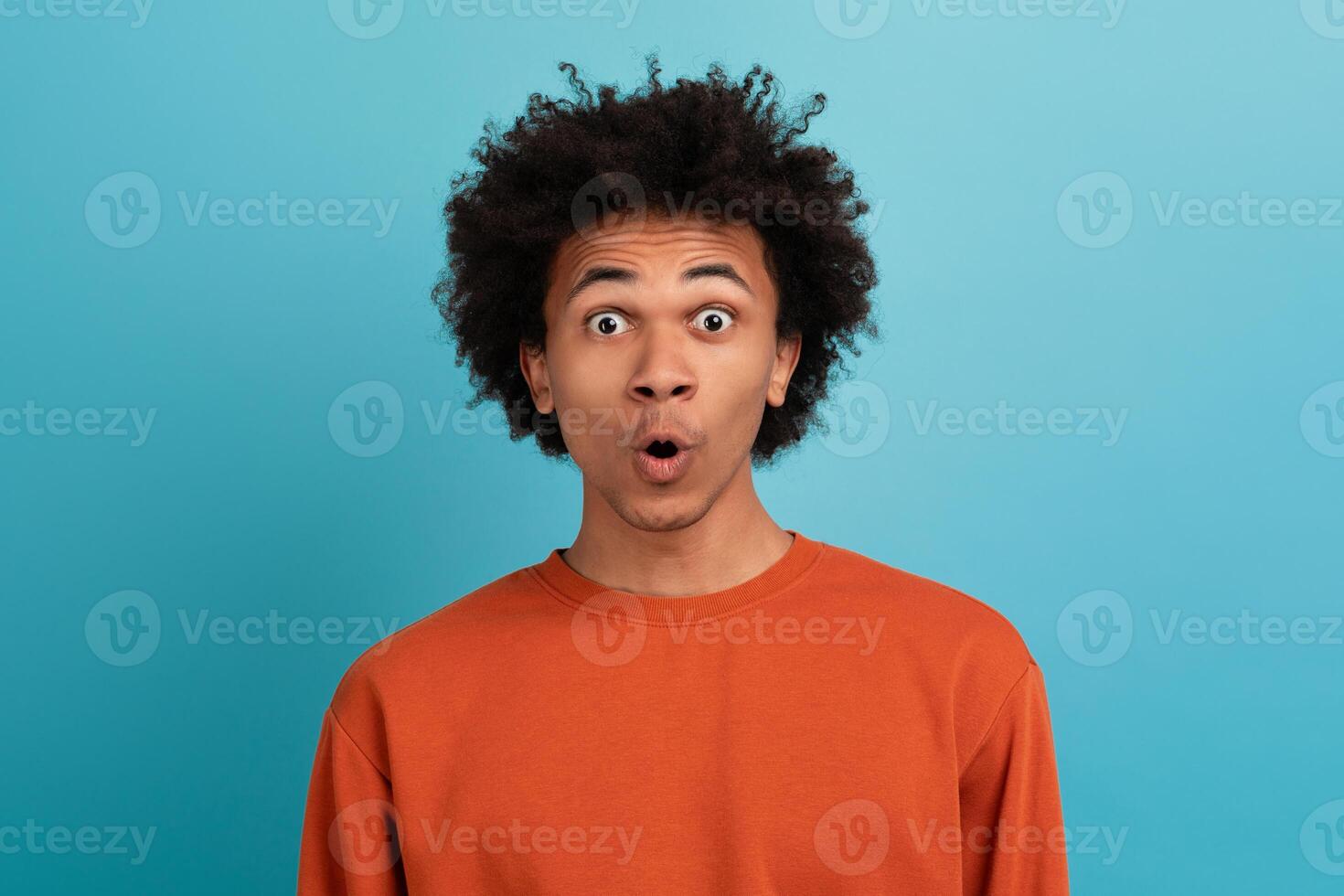 A young black man stands against a bright blue background, his curly hair framing his face. He shows a surprised expression, capturing a moment of joy and excitement. photo