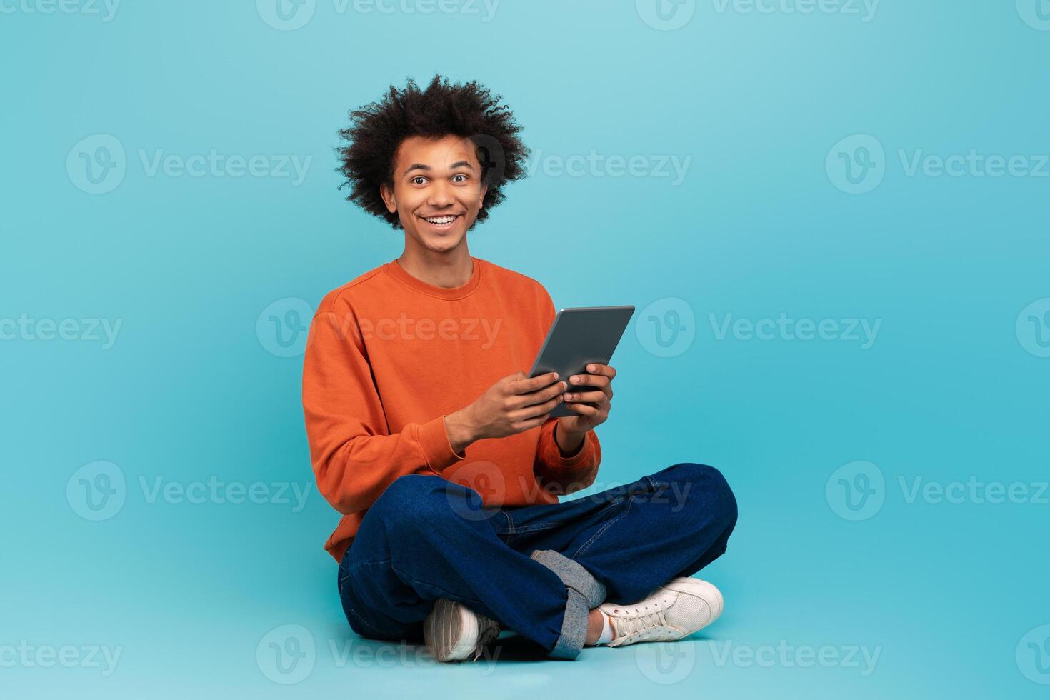 A young black man with curly hair sits comfortably on the floor, smiling while engaging with a tablet in his hands. The bright blue background adds a cheerful atmosphere. photo