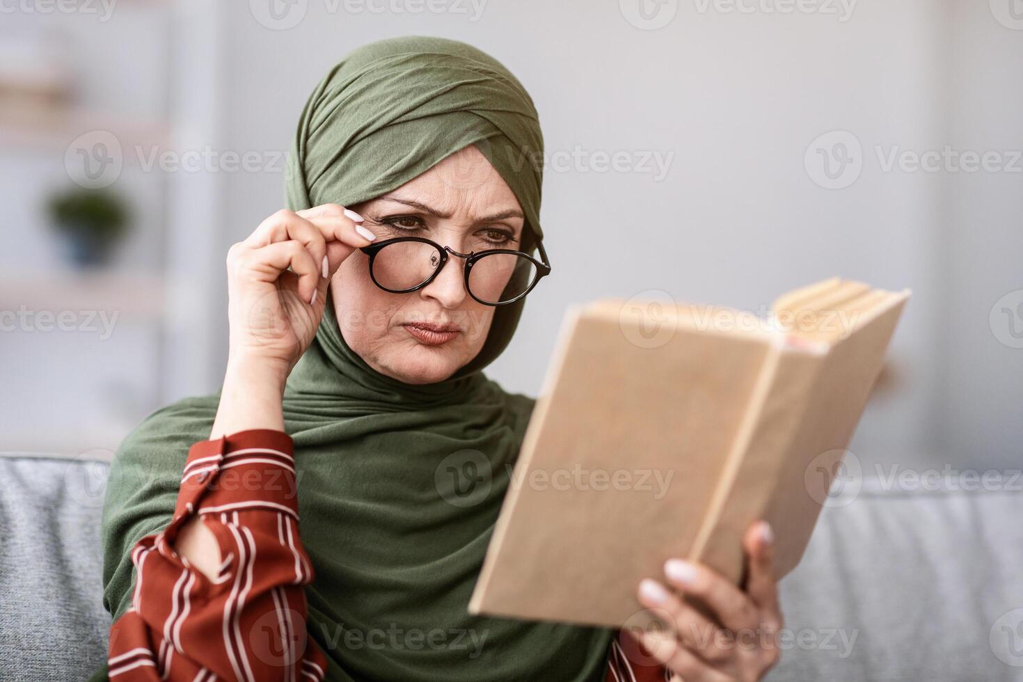 Senior Muslim Woman Having Poor Eyesight, Squinting Eyes While Reading Book Wearing Glasses At Home. Vision Healthcare Issue. Bad Vision And Eyes Health In Older Age Concept photo