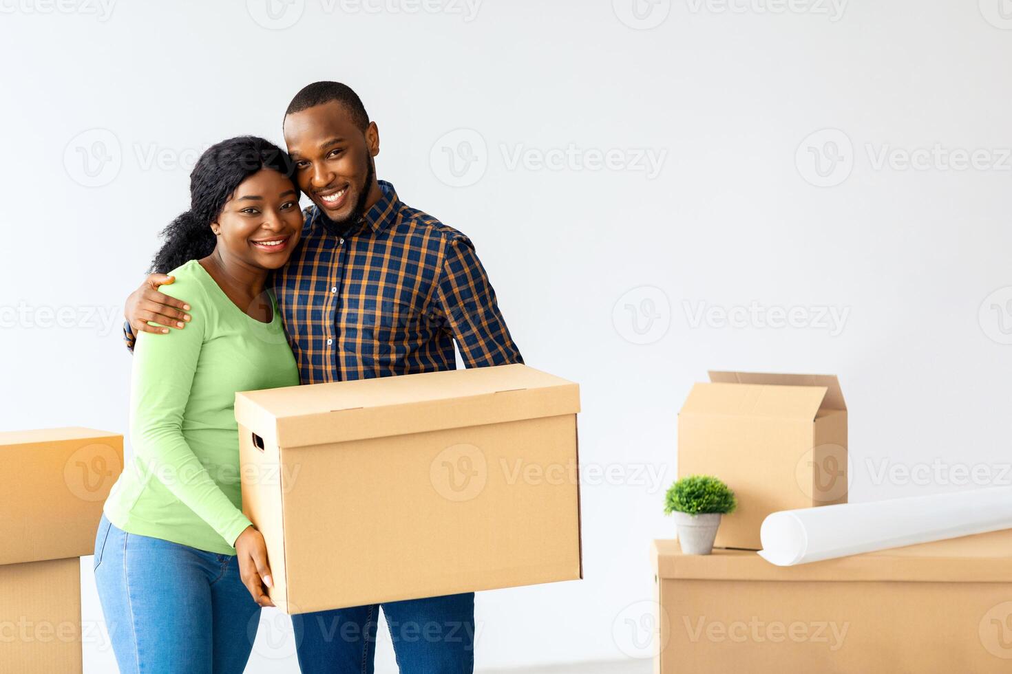 Couple Moving Concept. Happy Black Spouses Holding Cardboard Box And Smiling At Camera, Young Cheerful African American Wife And Husband Standing In New House, Celebrating Relocation, Copy Space photo