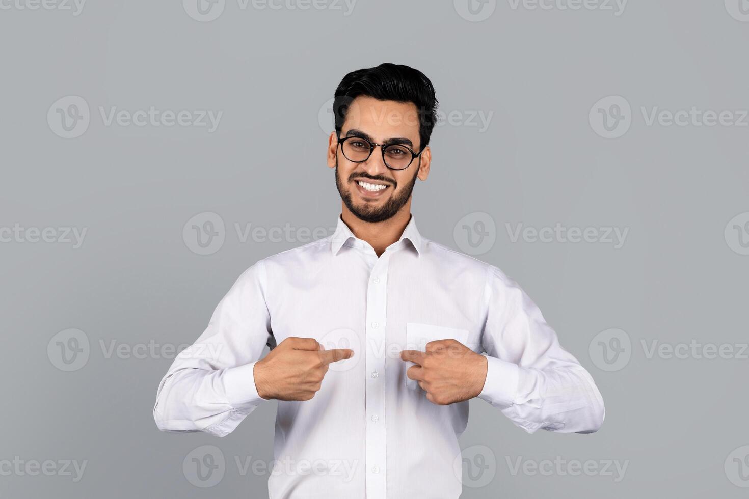 A Middle Eastern man in a white dress shirt smiles confidently while pointing to his chest. He stands against a neutral backdrop, conveying a modern, professional lifestyle. photo