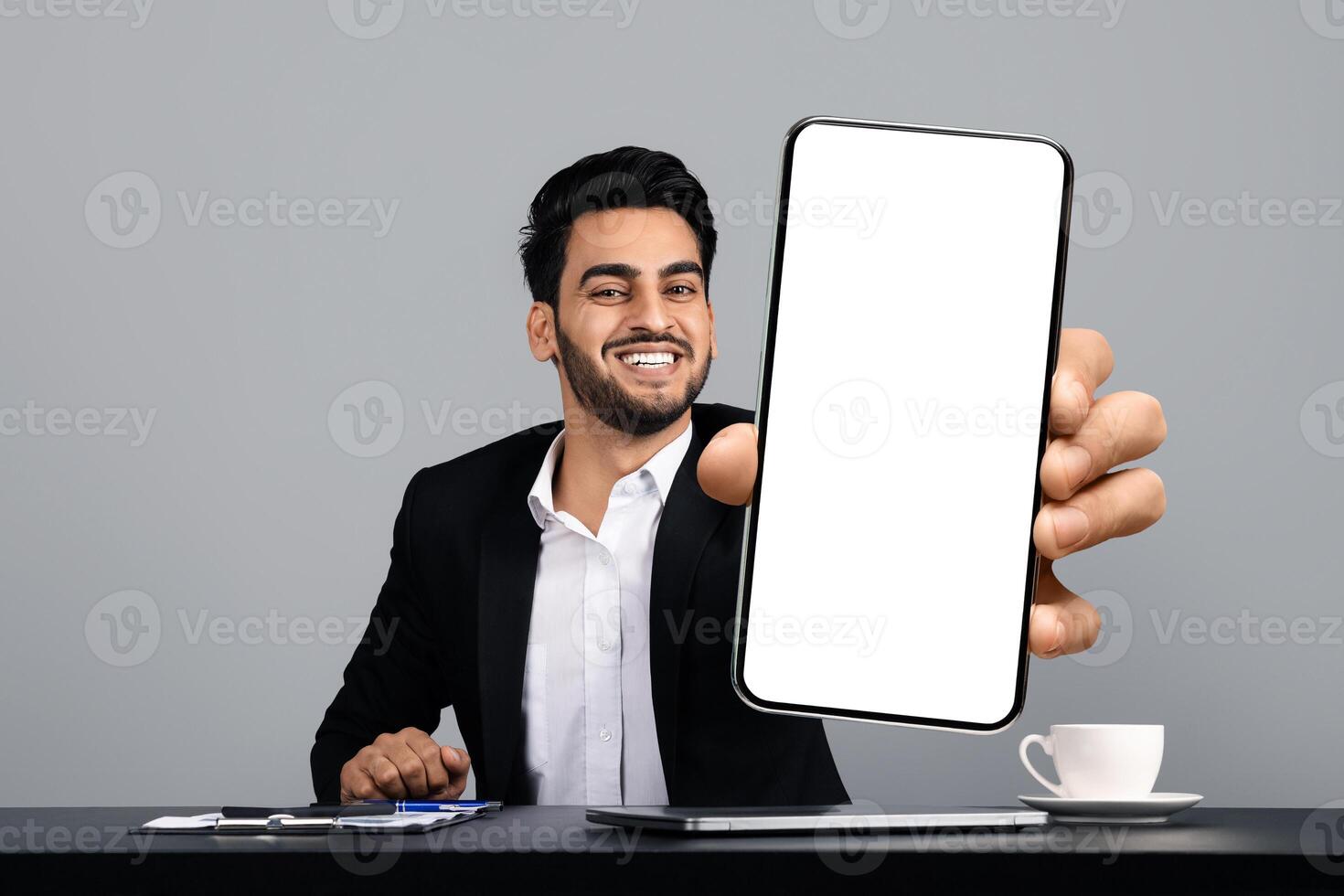 A young Arab entrepreneur sits at a desk in a modern studio, smiling confidently as he holds up a blank smartphone screen. He is dressed in a formal black suit, exuding a successful lifestyle. photo
