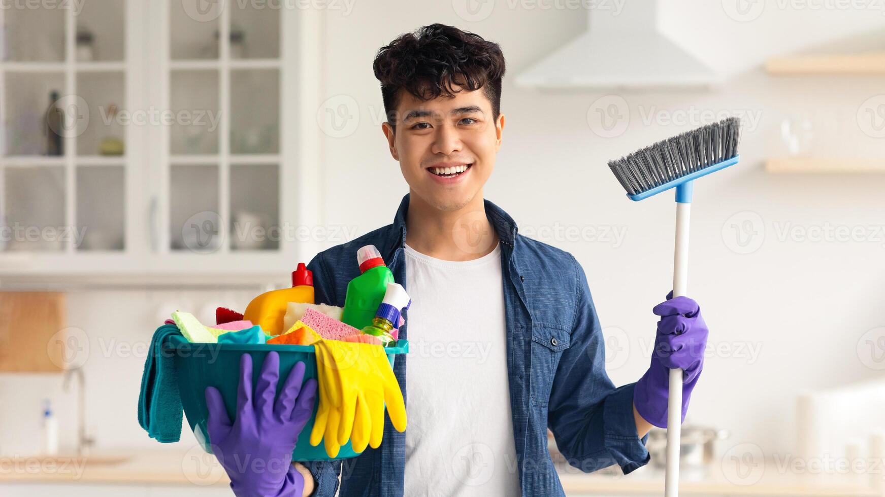 Smiling asian guy ready for cleaning apartment, holding mop and other cleaning tools, white cozy kitchen interior, copy space. Cheerful young man house-keeping on weekend photo