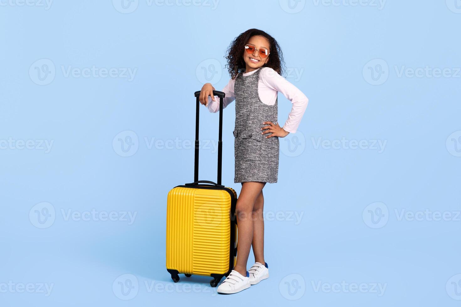 A cheerful black girl stands with a bright yellow suitcase, exuding excitement for an upcoming trip. Her stylish outfit and sunglasses reflect her playful preteen spirit in a vibrant setting. photo