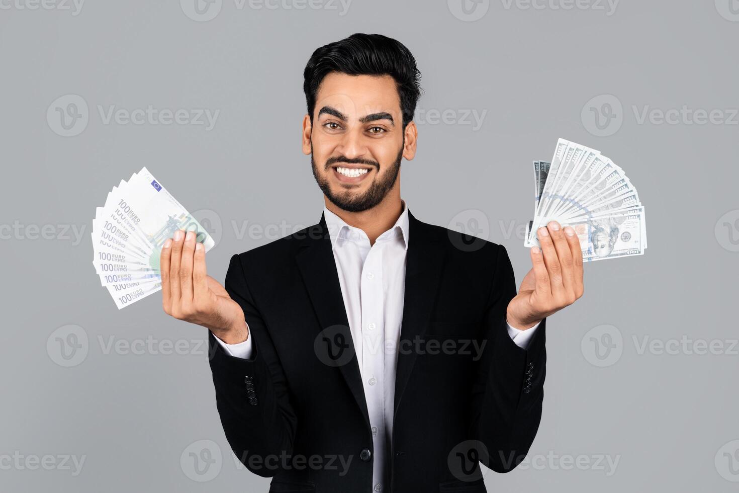 A confident Middle Eastern businessman displays bundles of cash in both hands while standing in a modern studio. He wears a professional outfit, reflecting a successful entrepreneurial lifestyle. photo