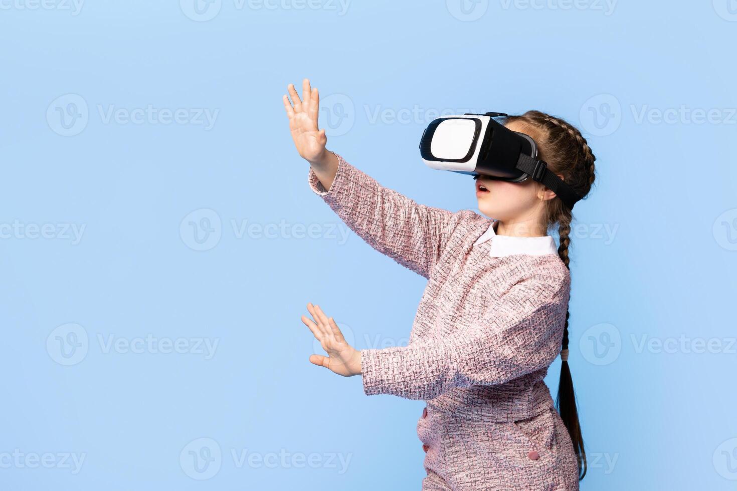 A joyful girl engages with virtual reality using a headset, demonstrating excitement and curiosity. She is dressed casually, immersed in her playful world, against a bright blue background. photo