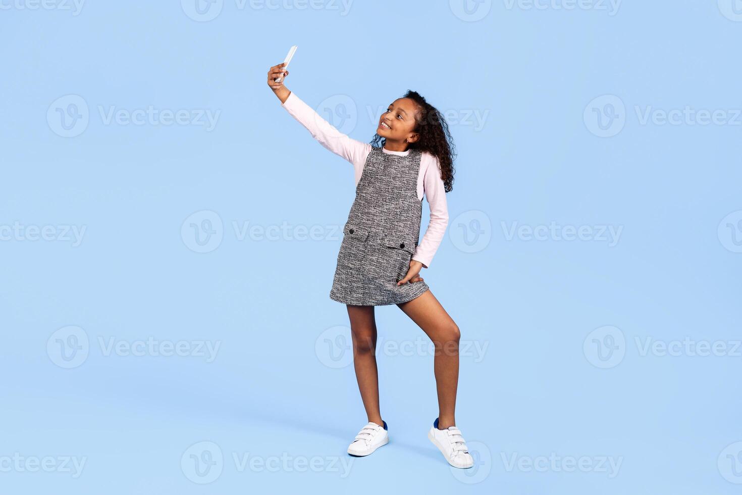 A joyful girl stands confidently while taking a selfie. Her cheerful expression and playful pose embody the spirit of childhood and fun. The vibrant blue background emphasizes her happiness. photo