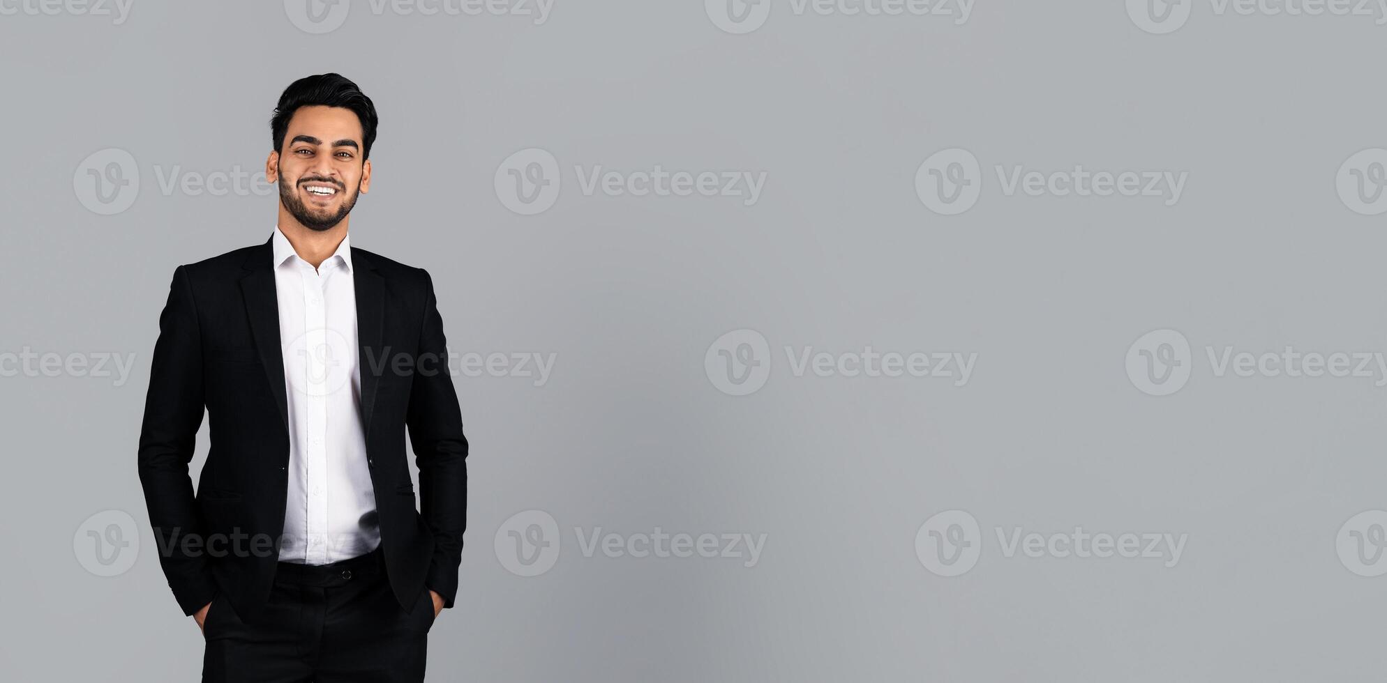 A Middle Eastern entrepreneur stands confidently in a studio, dressed in a formal black suit with a white shirt. His smile reflects success and professionalism in the corporate world, copy space photo