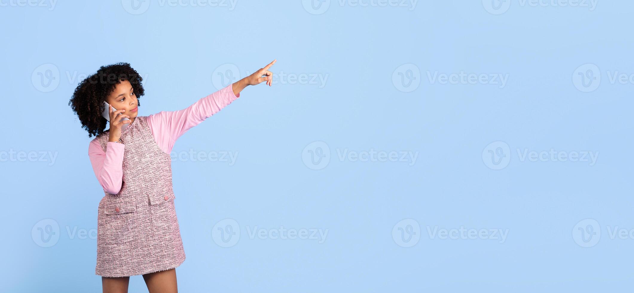 A joyful preteen girl engages in a phone conversation, smiling and pointing at copy space as if sharing an exciting story. She stands alone against a bright blue backdrop photo