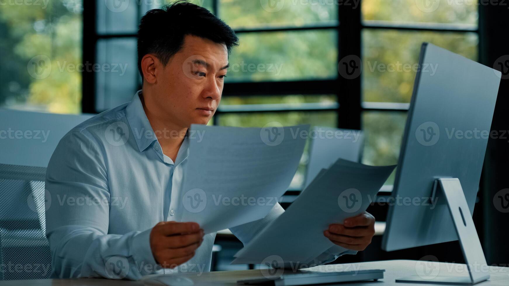 Tired unhappy Asian man worker manager sit at office doing routine paperwork read contract documents budget invoice pay bills online analyze business plan check documentation professional paper audit photo