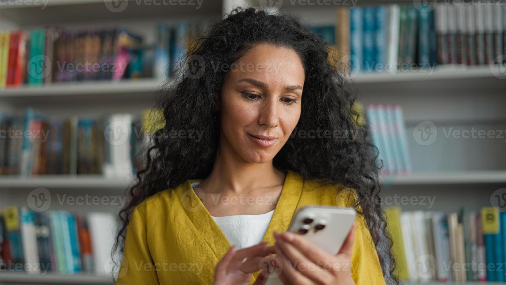 Latin Hispanic Arab woman student mobile phone user smartphone texting sms chatting gadget library university high school college networking smile female girl laugh technology browsing e-learning web photo
