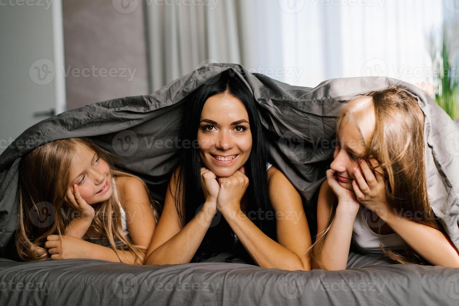 Mom and two daughters are lying on the bed under a blanket. The brunette mother looks at the camera, and the girls look at her and smile photo
