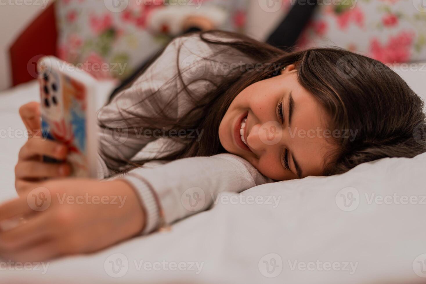 A smiling teenage girl using a phone while comfortable lying on the bed at home. photo