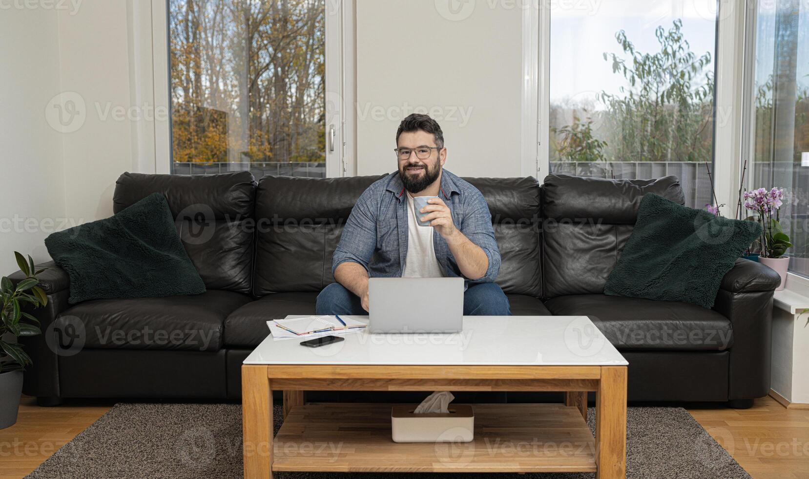 Happy man drinking coffee using a laptop sitting on comfortable couch at home in living room. photo