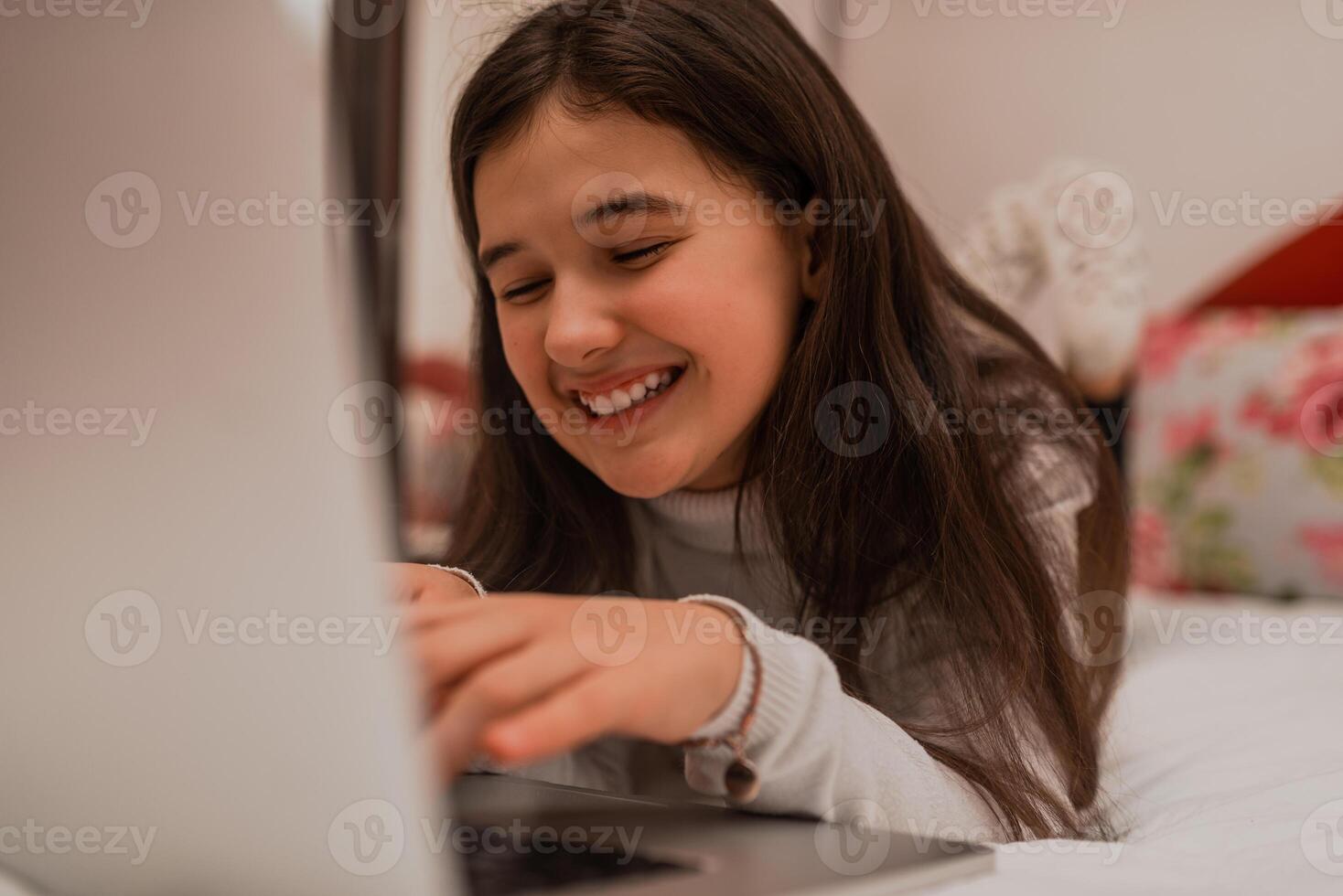 A smiling teenage girl using a laptop while comfortable lying on the bed at home. photo