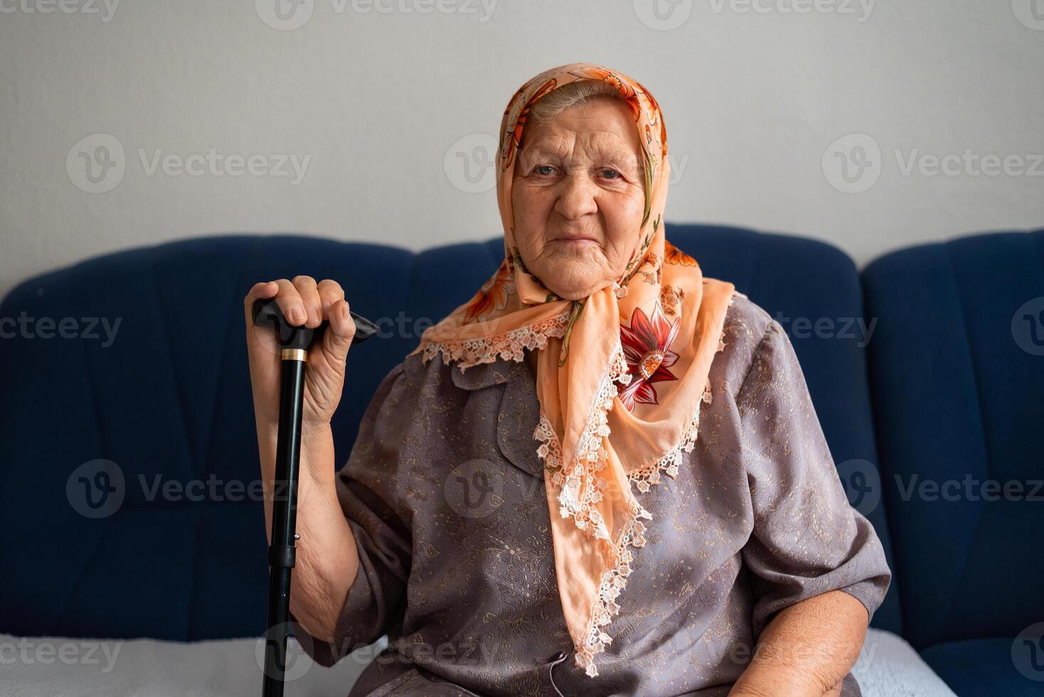 Portrait of an old sad Muslim woman with her walking stick at home wearing traditional hijab, looking at the camera. photo