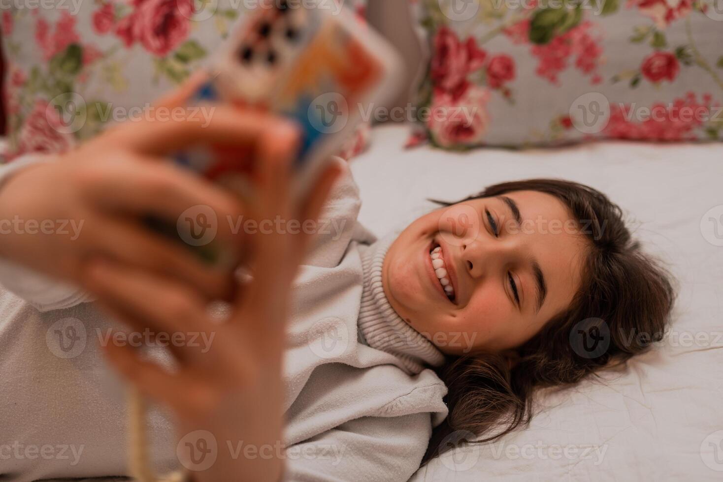 A smiling teenage girl using a phone while comfortable lying on the bed at home. photo