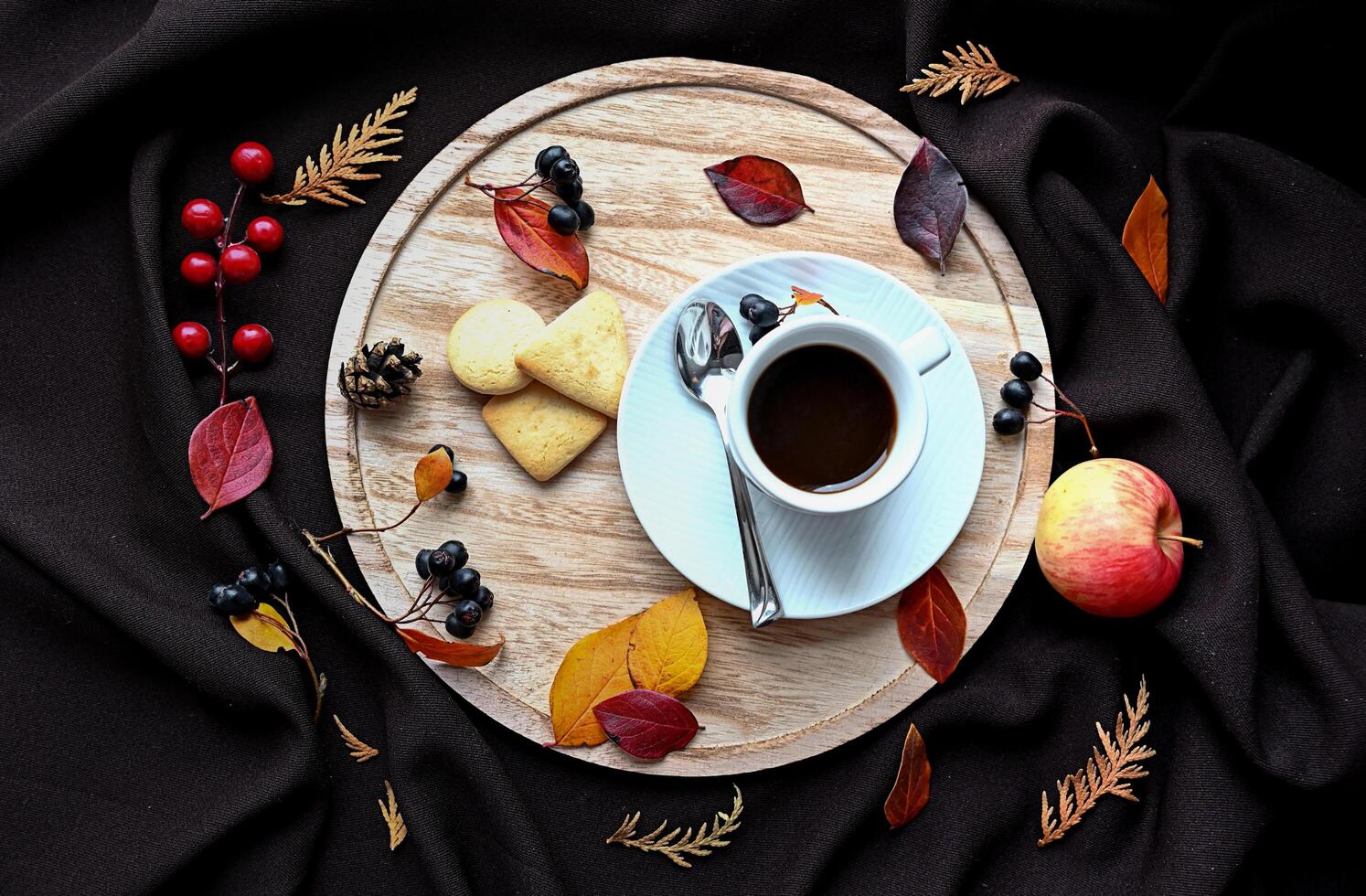 There is a cup of coffee, a saucer, a spoon, three cookies, an apple, and various autumn leaves on the tray. photo