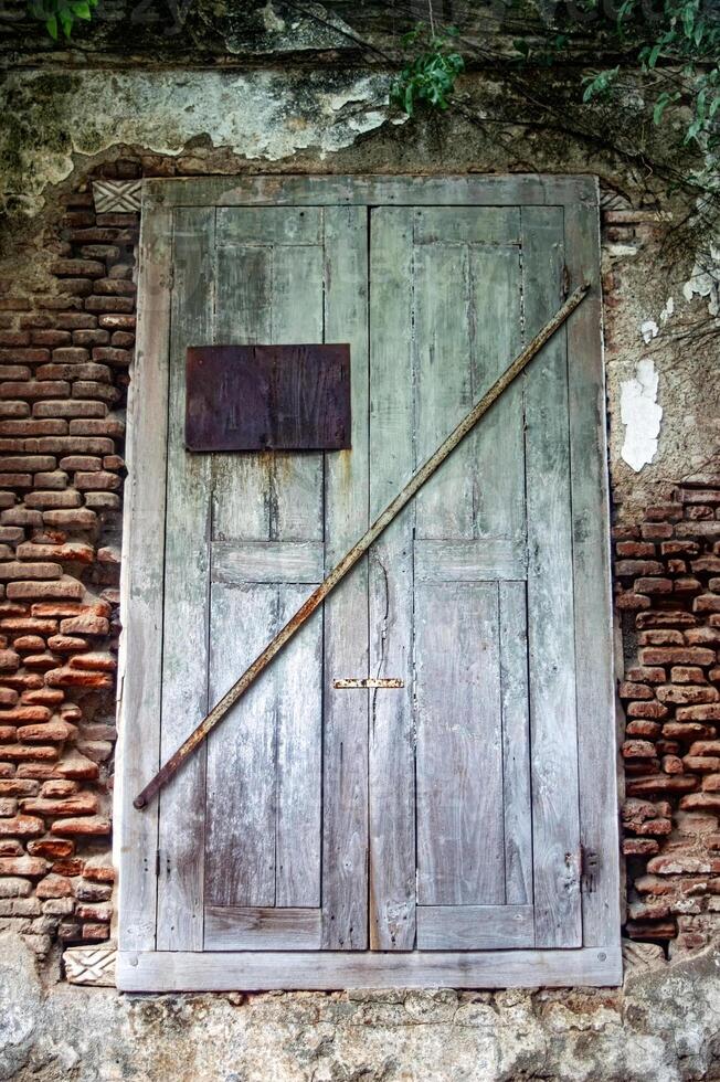 An old wooden door with peeling paint, secured by a rusty metal bar, set in a weathered brick wall with creeping vines photo