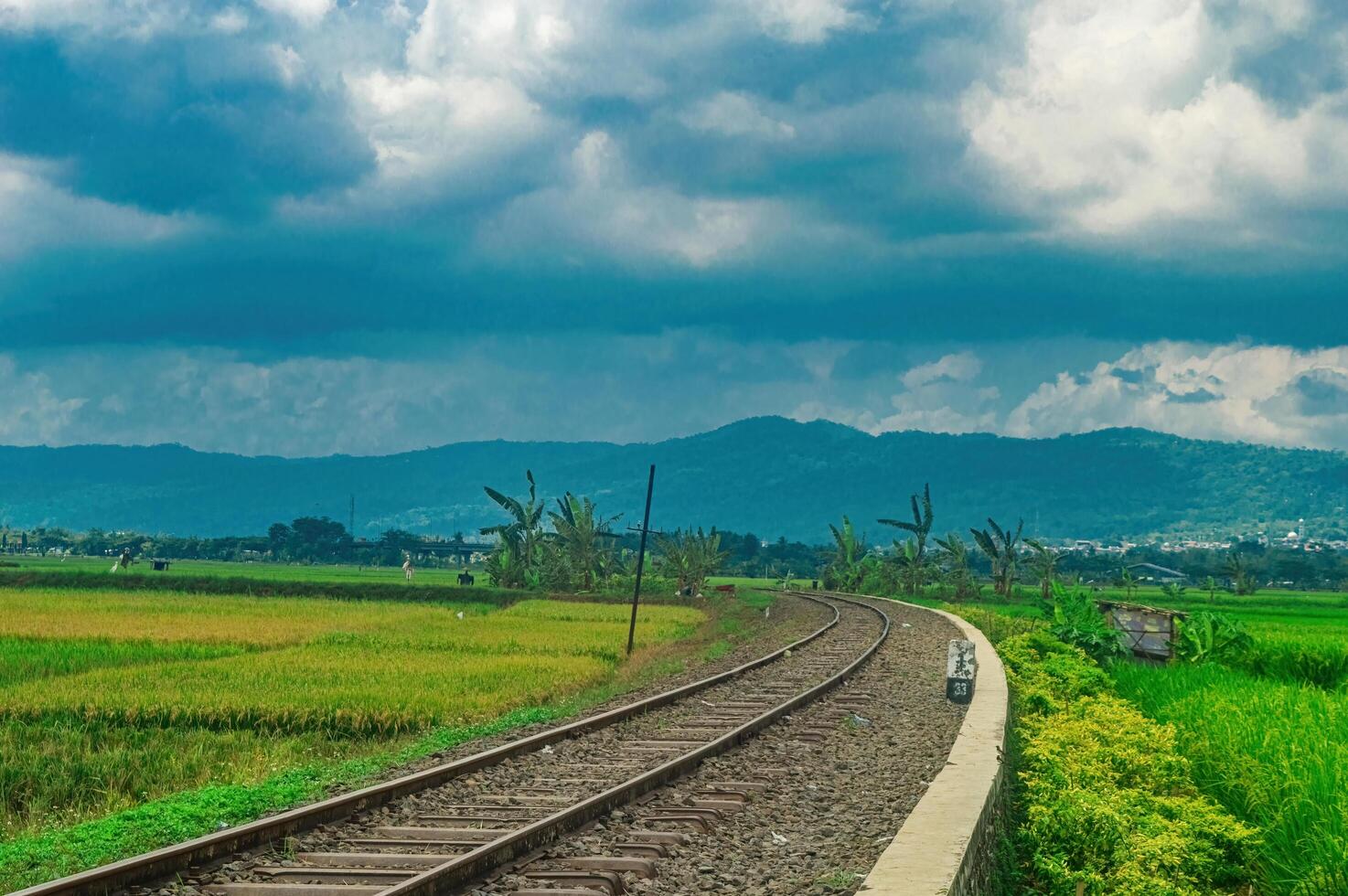 Scenic railway track curving through lush green fields with a backdrop of majestic mountains and dramatic clouds, perfect for travel and nature enthusiasts photo
