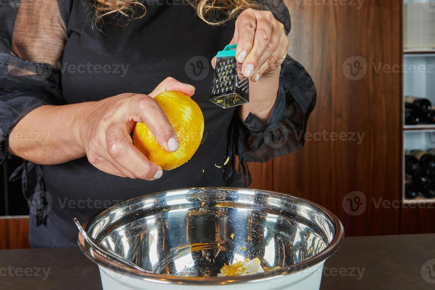 A person zesting an orange using a small grater over a metallic mixing bowl, adding a fresh citrus flavor to the recipe. photo