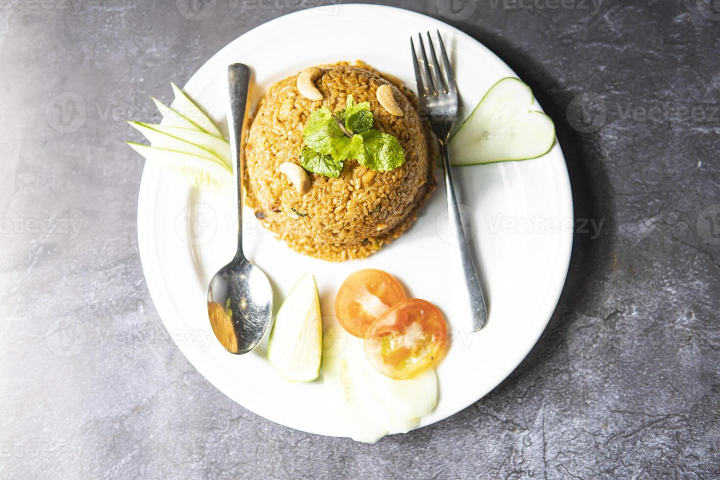 Chicken Dum Biryani topped with cashew nut, mint leave, cucumber, tomato, fork and knife served in plate isolate on grey background top view of indian, bangladeshi and pakistani food photo