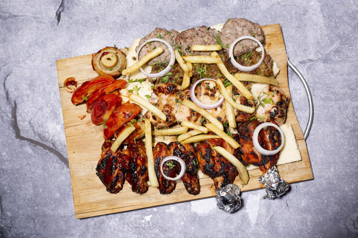 Mix Grill Bbq Platter including Chicken Tangdi kabab, Chicken Reshmi Tikka Kabab, Beef Bihari Kebab, Turkish shish kebab, Fries, Tomato, Onion and Mushroom served on wooden board top view photo
