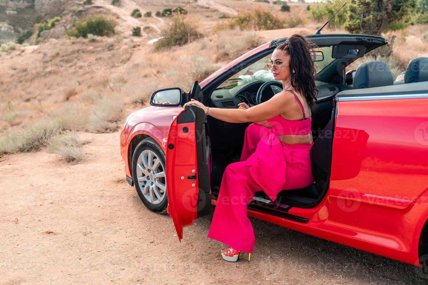 A woman in a pink outfit is sitting in a red convertible car. The car is parked on a dirt road, and the woman is enjoying the ride. photo