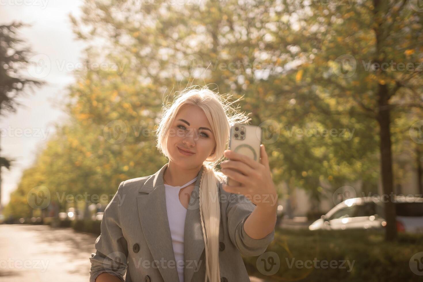 A woman is taking a picture of herself with her cell phone. She is wearing a gray jacket and scarf. The scene is set in a city with trees and cars in the background. photo
