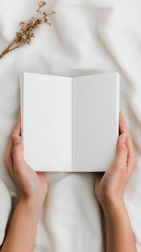 Open blank notebook held by hands on soft fabric with dried flowers nearby in natural light photo