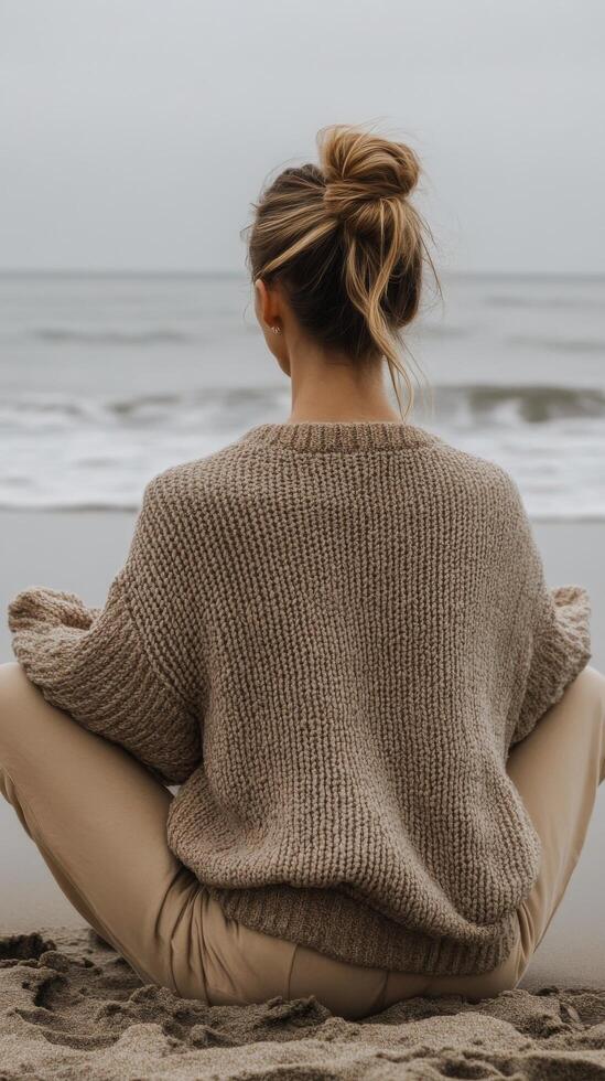 A woman with her hair tied back sits cross legged on the sandy beach, wearing a cozy sweater photo