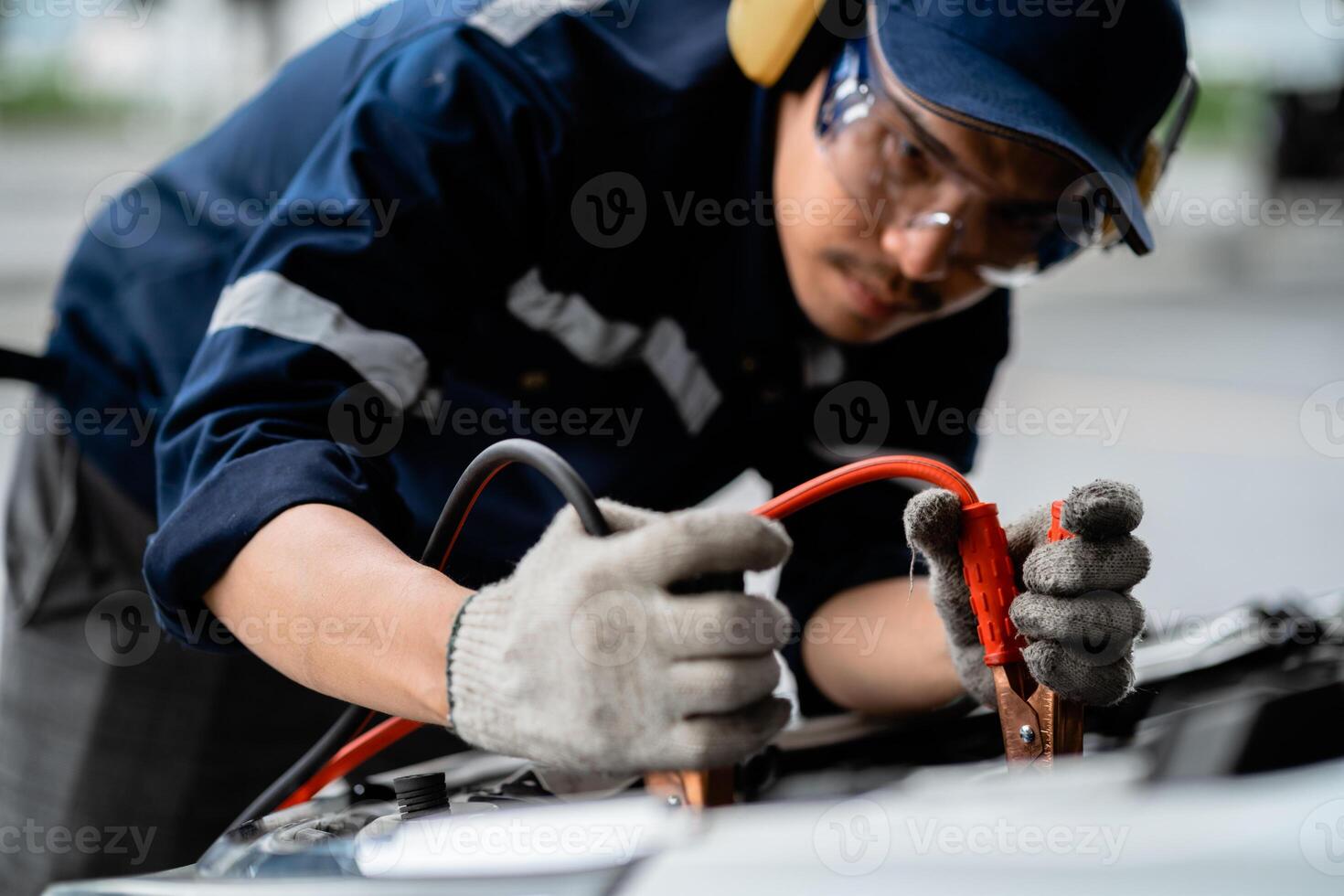 A man in a blue shirt and goggles is working on a car. He is using a jumper cable to jump start the car photo