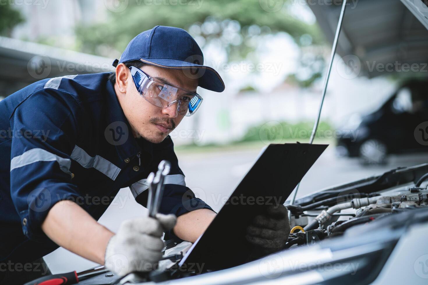A mechanic is working on a car engine, looking at a clipboard with a list of repairs. The mechanic is wearing a blue shirt and a blue hat photo