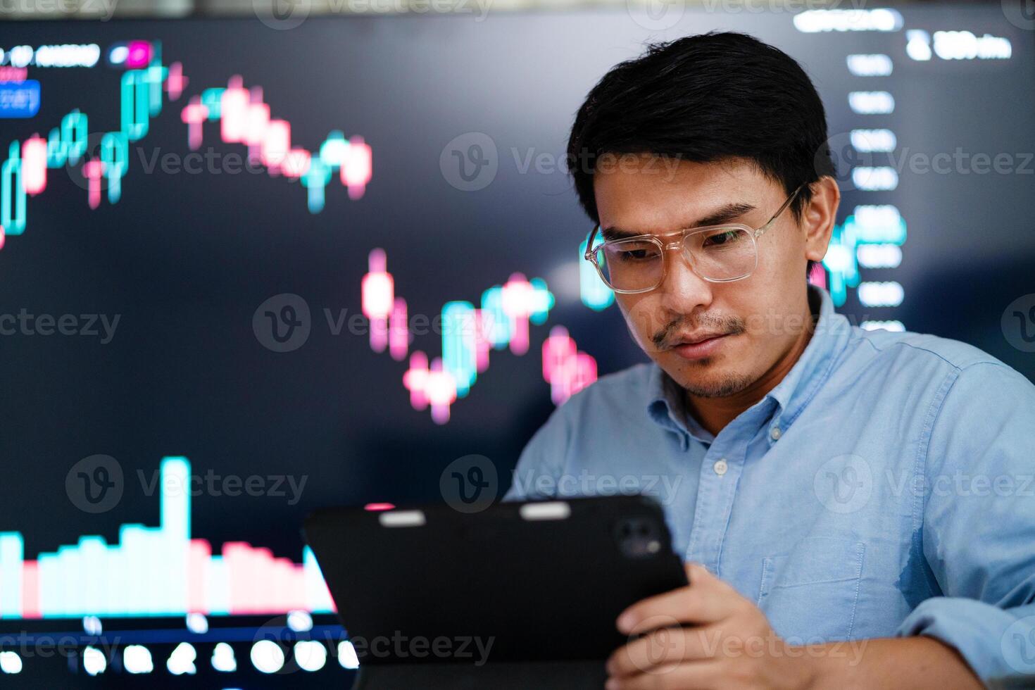 A man is looking at a computer screen with a lot of numbers and graphs. He is wearing glasses and holding a tablet photo