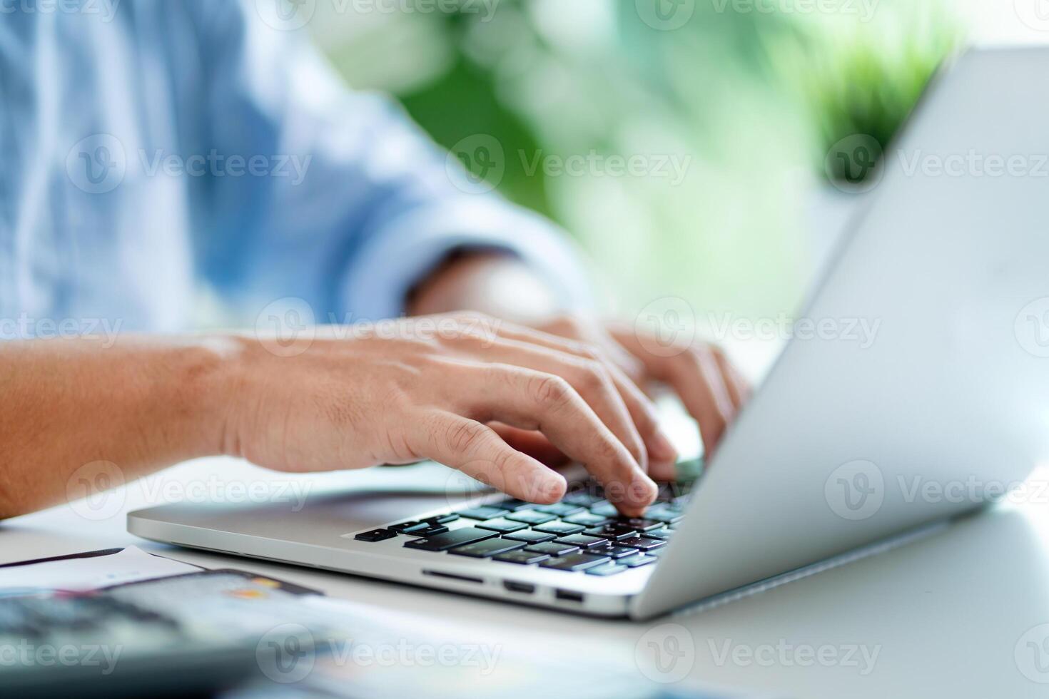 A man is typing on a laptop computer. He is wearing a blue shirt. The laptop is open and the keyboard is visible photo