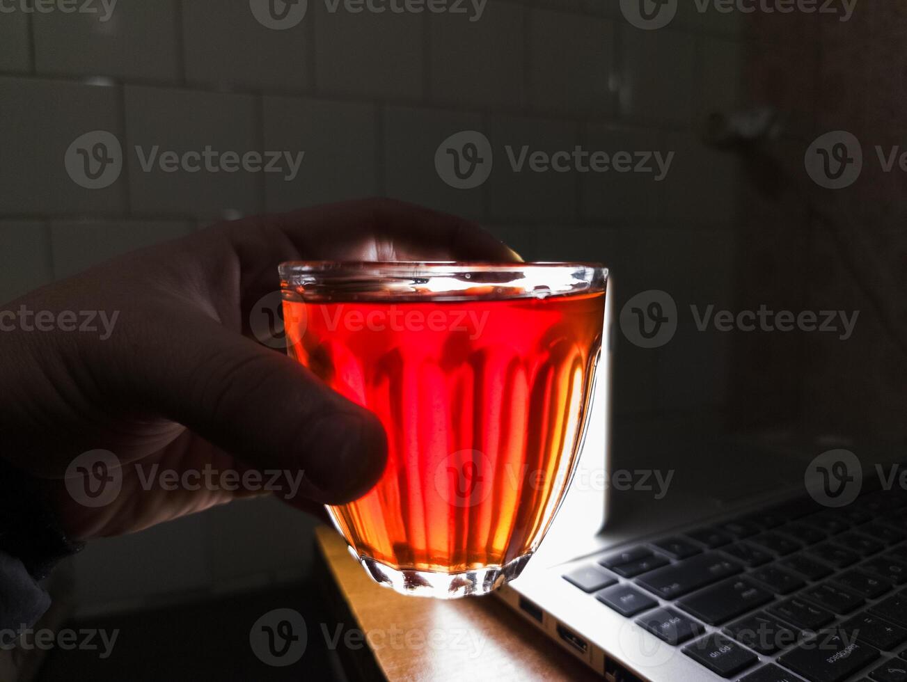 A person holding a glass of red liquid in front of a laptop computer photo