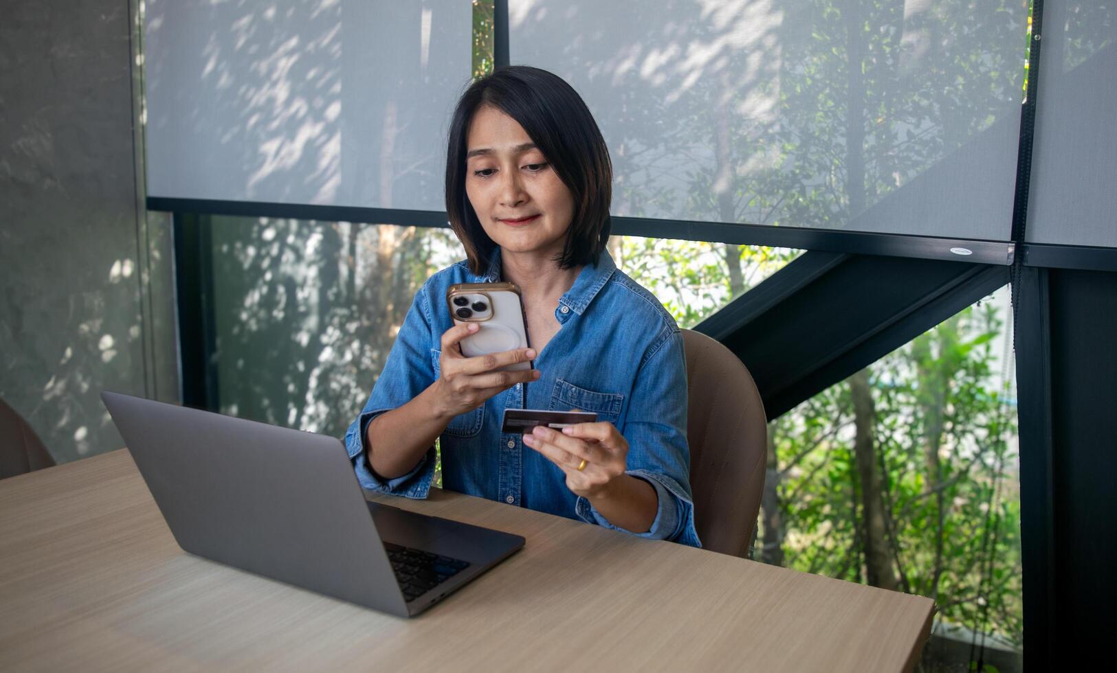 Asian woman sitting on chair holding mobile phone credit card in hand and looking at laptop for shopping online, internet banking and payment success, relaxing lifestyles concepts photo