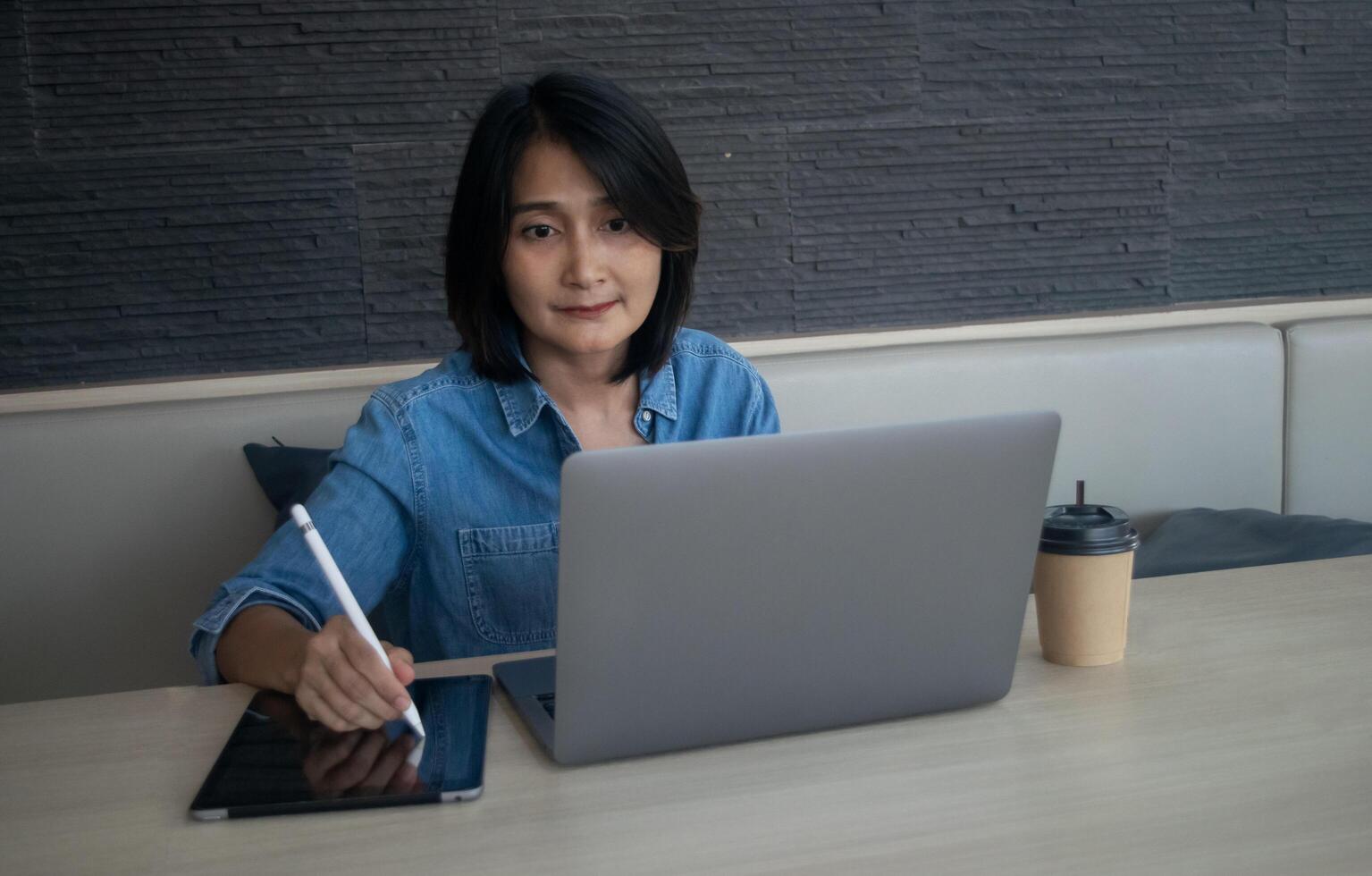 Asian woman hand holding pen and using digital tablet, her looking at working laptop analysis financial reports and coffee cup put on desk for drinking, online learning concepts photo