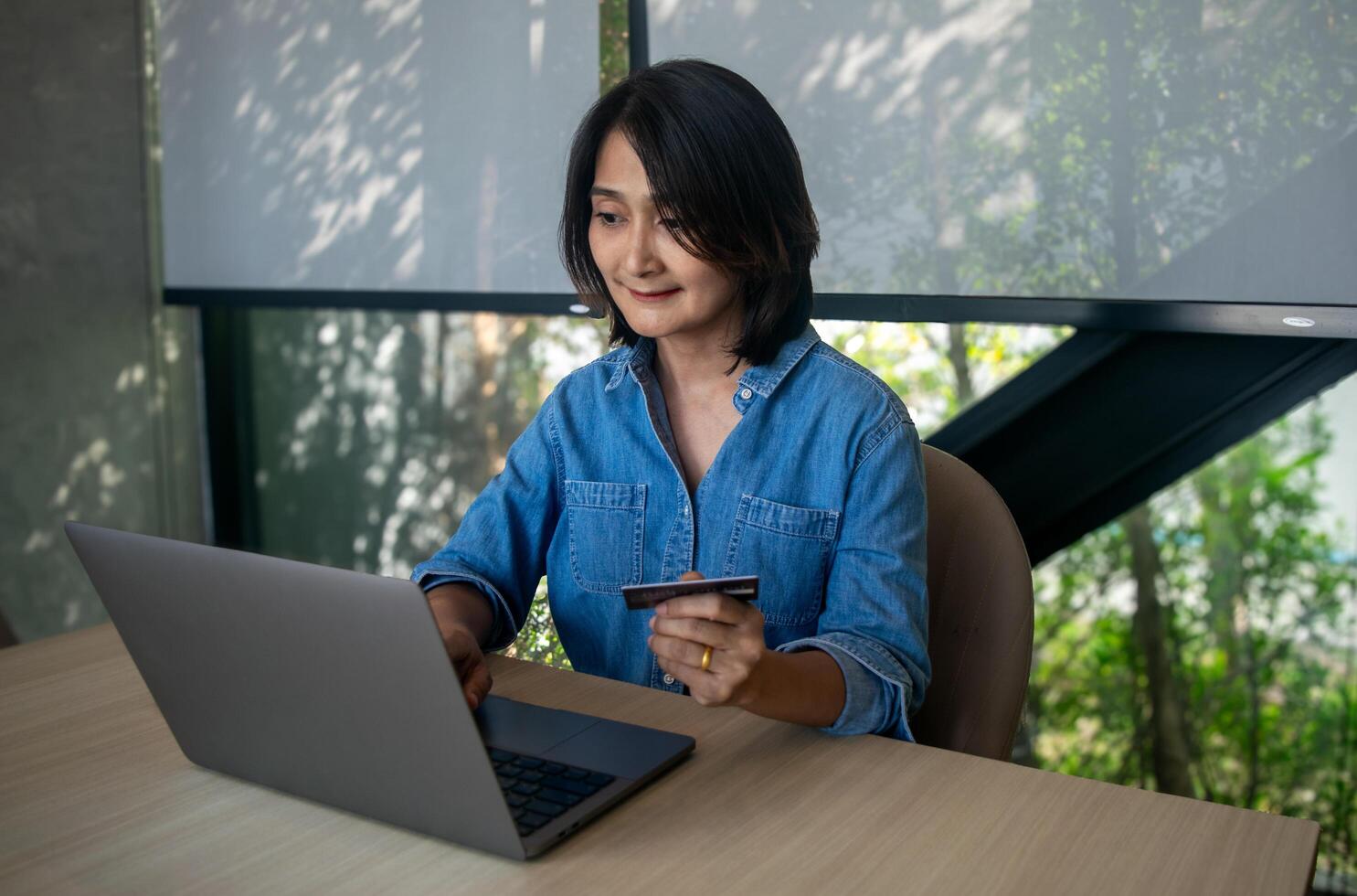 Asian woman sitting on chair holding credit card in hand for online shopping using laptop and buying order on website paying marking purchase online store internet banking finance technology photo