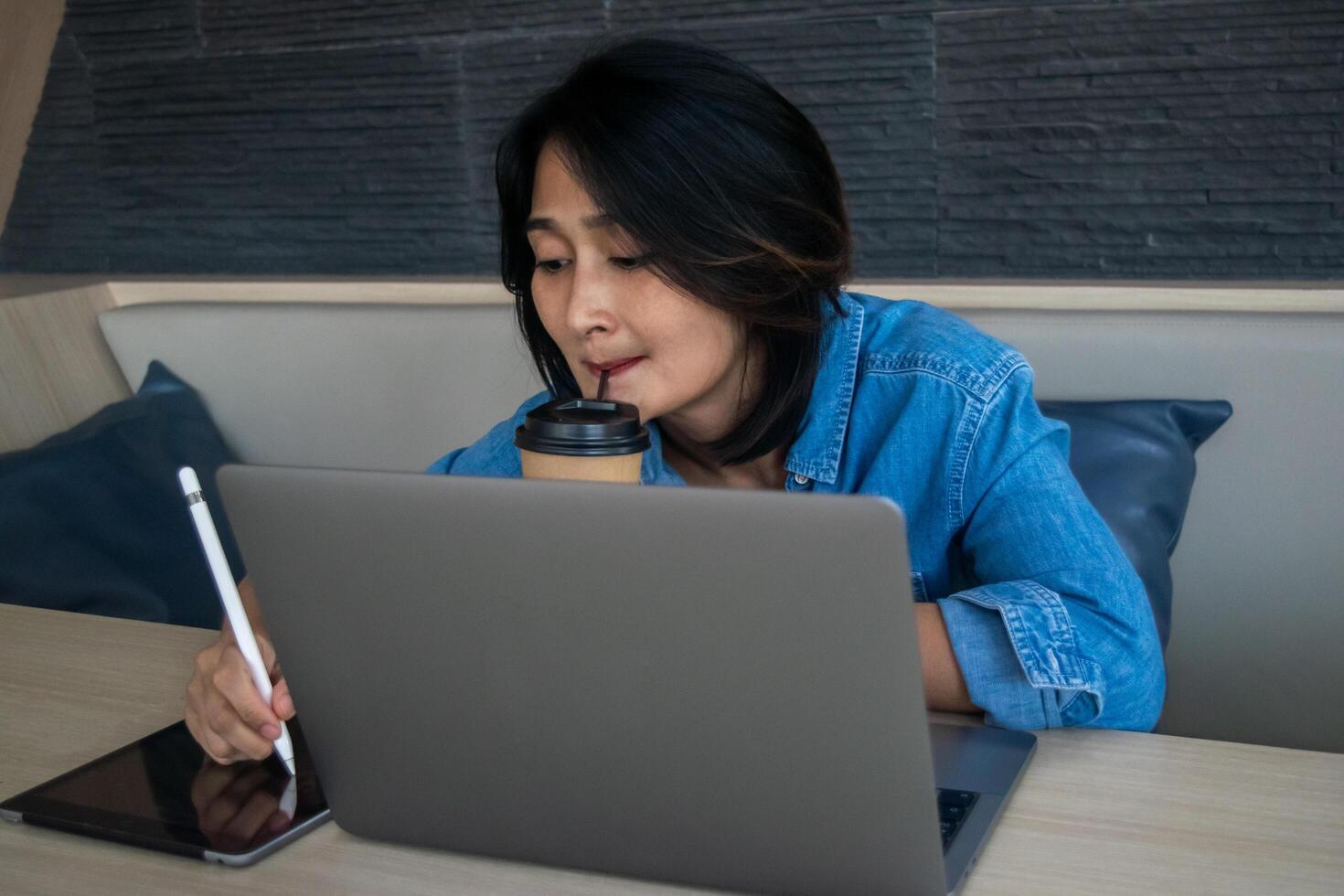 Asian woman hand holding pen and using digital tablet, her looking at working laptop analysis financial reports and drinking coffee cup, online learning concepts photo