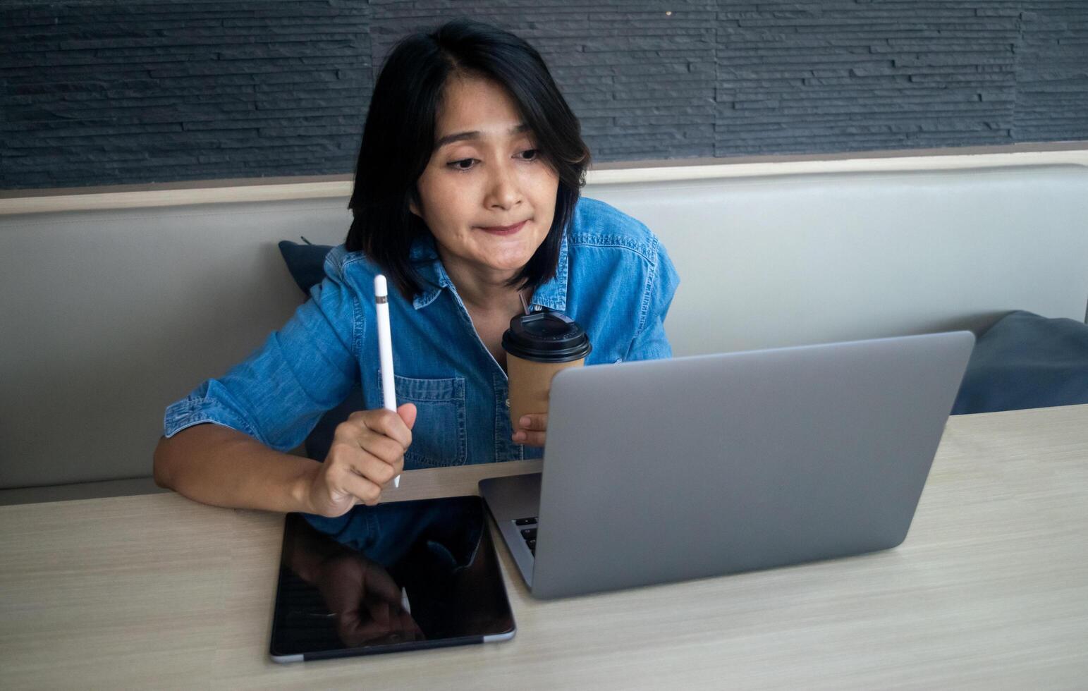 Asian woman hand holding pen and using digital tablet, her looking at working laptop analysis financial reports and drinking coffee cup, online learning concepts photo