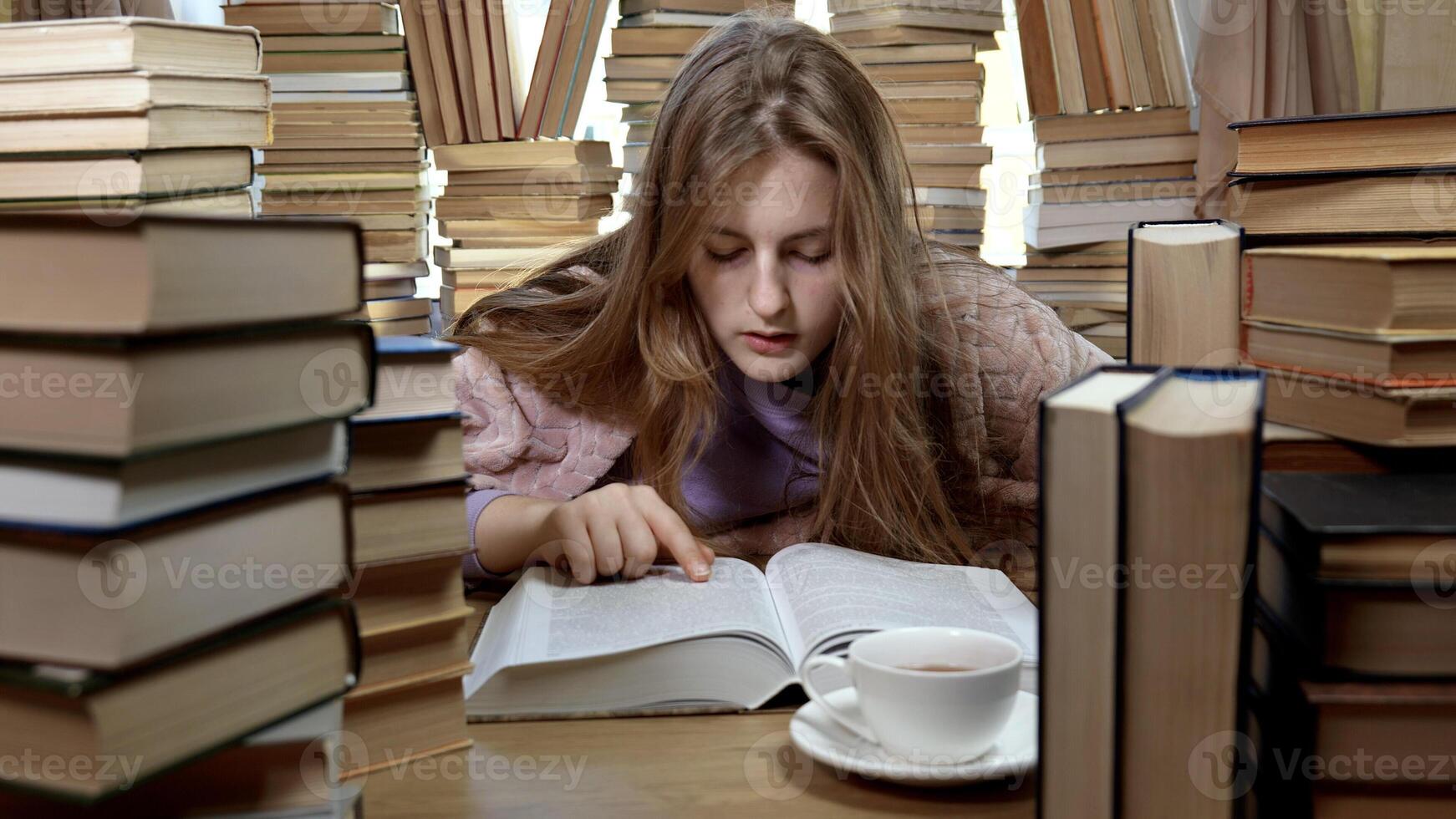 A girl reads a book in the library. Stacks of books and cup of tea on table. Concept of Reading and Education. photo