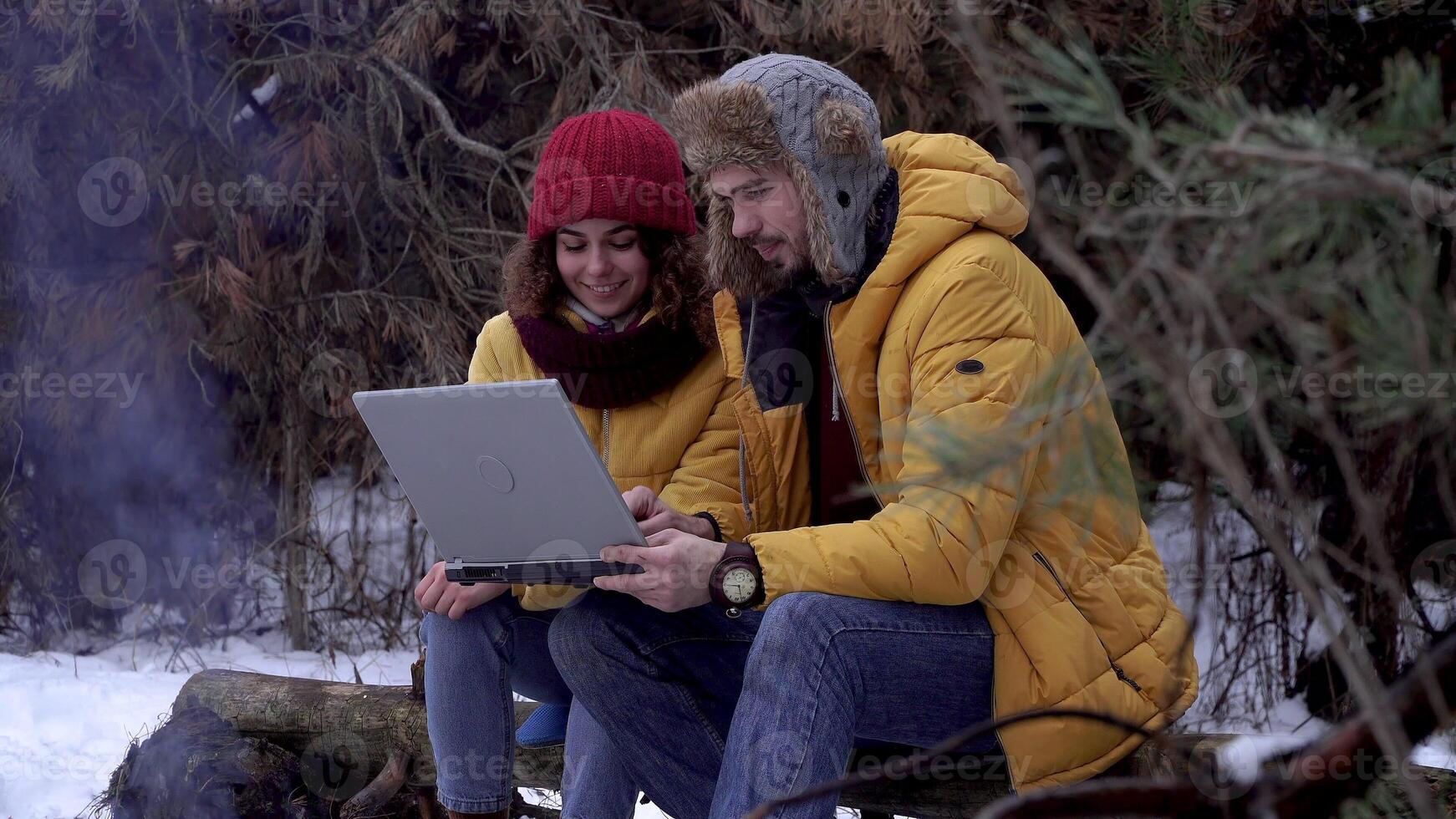 Young man and woman with a laptop near a campfire in the winter forest. Tourists travel together. The guys are smiling and chatting. photo